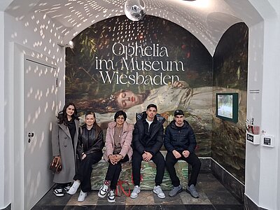 Fünf Personen sitzen auf einer Bank vor einer großen Wandgestaltung mit dem Gemälde „Ophelia“ und dem Text „Ophelia im Museum Wiesbaden“ in einem weiß gewölbten Raum mit Spiegelkugel an der Decke.