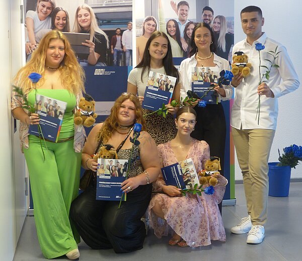 Zu sehen sind fünf junge Frauen in verschieden farbigen Kleidern, die einen Blumenstrauß und ihr Abschlusszeugnis der Euro Akademie in der Handhalten. Rechts daneben steht ein junger Mann mit weißem Hemd, der diese Dinge ebenfalls in der Hand hält.