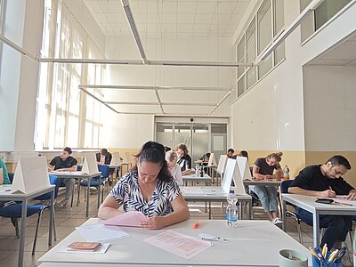 Die Prüflinge sitzen in einem großen, hellen Raum mit hoher Decke an Tischen, es sind Trenner als Sichtschutz zwischen ihnen aufgestallt, alle schrieben konzentriert ihre Antworten auf, vor ihnen auf dem Tisch liegen Schreibuntensilien und etwas zu trinken. 