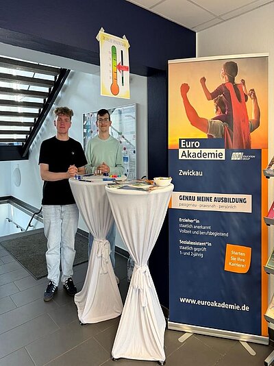 Zwei junge Männer stehen hinter einem Informationstisch der Euro Akademie Zwickau. Der Tisch ist mit Informationsmaterial und einem Roll-up-Banner der Euro Akademie versehen, auf dem „Genau meine Ausbildung“ und die Website „www.euroakademie.de“ zu lesen sind. Im Hintergrund hängt ein Thermometer-Poster mit einer Spendenanzeige.