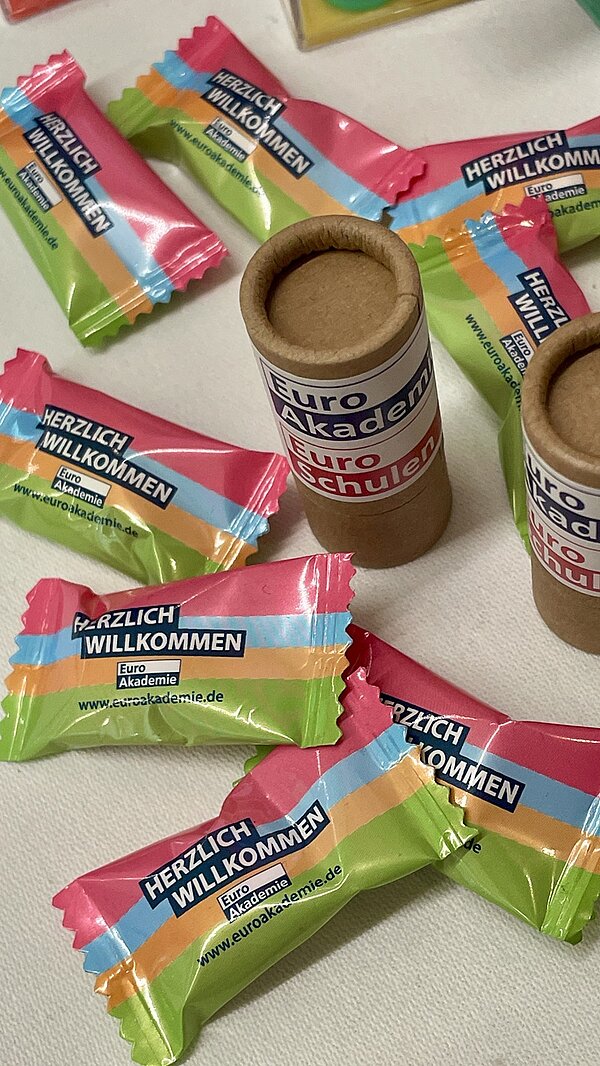 Mehrere Bonbons mit einer auffälligen, farbigen Verpackung liegen auf einer hellen Oberfläche. Die Verpackung ist in den Farben Pink, Grün und Gelb gestaltet und trägt den Aufdruck 'HERZLICH WILLKOMMEN' sowie ein Logo mit der Aufschrift 'Euro Akademie'. Daneben stehen zwei zylinderförmige Behälter aus braunem Karton, die mit einem weißen Etikett beklebt sind. Auf dem Etikett steht 'Euro Akademie' und 'Euroschulen'.