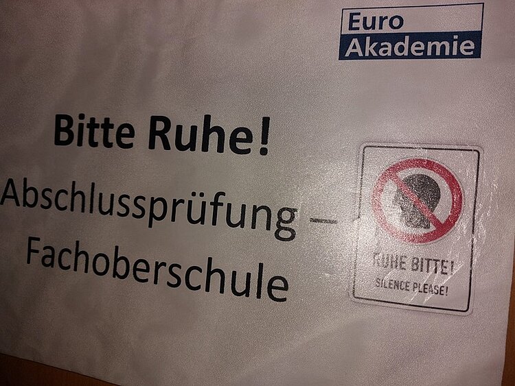 Ein rechteckiges Schild mit dem Logo und Schriftzug der Euro Akademie oben rechts. Darunter steht in großen Buchstaben: 'Bitte Ruhe! Abschlussprüfung – Fachoberschule'. Rechts daneben befindet sich ein einzelnes Piktogramm mit einem durchgestrichenen Lautsprechersymbol und dem Text 'RUHE BITTE! SILENCE PLEASE!'. Der Hintergrund ist hell, die Schrift schwarz.