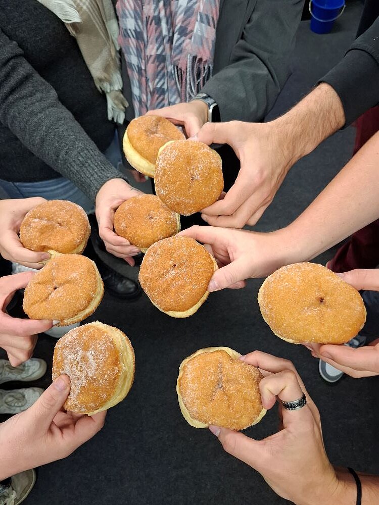 Mehrere Personen stehen im Kreis und halten jeweils einen Berliner beziehungsweise Pfannkuchen in den Händen, die sie in die Mitte strecken. Die goldbraunen, zuckrigen Gebäckstücke symbolisieren den gemeinsamen Start in die Faschingszeit.