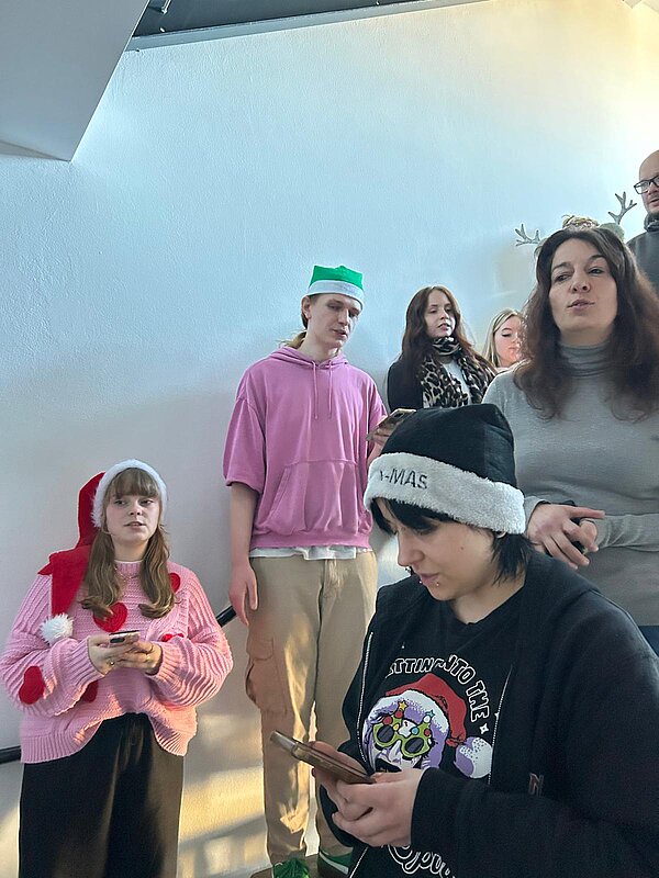 Mehrere junge Menschen stehen auf einem Treppenabsatz, einige tragen Weihnachtsmützen und lesen Liedtexte von ihren Smartphones.