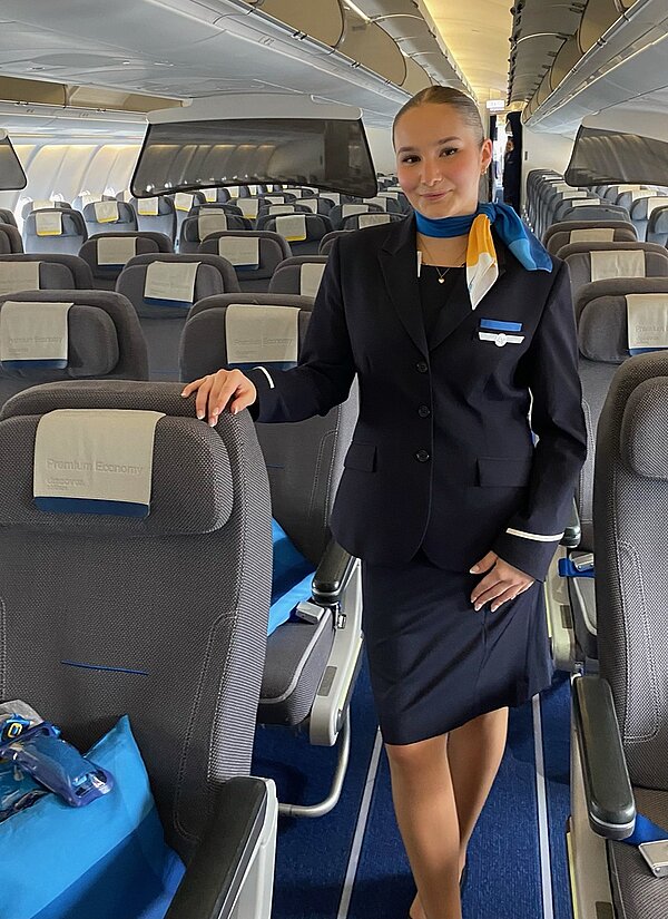 Alumni im Gespräch: Interview mit Lea Findeis Die Euro Akademie Absolventin Lea Findeis steht in ihrer Berufskleidung als Flugbegleiterin im Gang eines leeren Flugzeuges und lächelt für ein Foto.