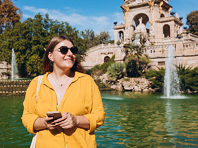 Eine lächelnde junge Frau mit Sonnenbrille und gelber Jacke steht vor einem großen, historischen Brunnen mit Statuen und Wasserfontänen. Sie hält ein Smartphone in der Hand und scheint die Umgebung zu genießen.