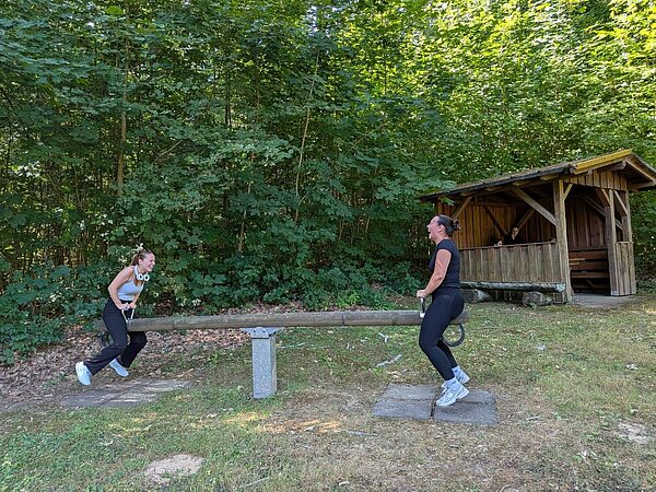 Zwei Personen sitzen auf einer Wippe im Wald und lachen. Eine Person davon trägt weiße Kopfhörer um den Hals. Im Hintergrund ist eine Rasthütte aus Holz, in der eine weitere Person sitzt sowie dichte Bäume zu sehen.