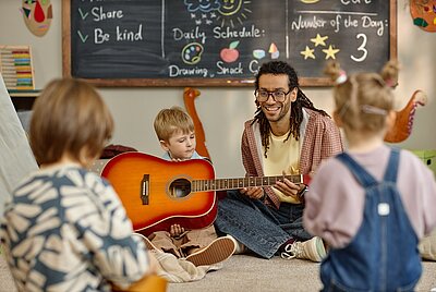Ein Mann mit Brille und langen, lockigen Haaren sitzt auf dem Boden in einem Raum mit Kindern. Ein Junge hält eine rote Akustikgitarre, während zwei weitere Kinder im Vordergrund stehen. Im Hintergrund ist eine Tafel mit handgeschriebenen Notizen und Zeichnungen zu sehen. Die Szene wirkt freundlich und entspannt.