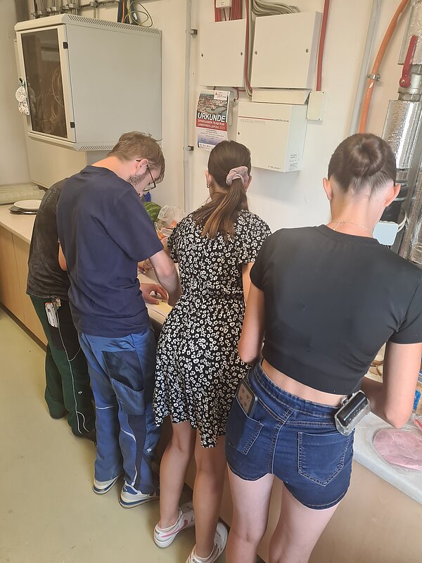 DRK Werkstätten Geithain - Aktionstag gesunde Ernährung Zu sehen sind zwei Junge Frauen und ein Mann, die vor einem Werkstatttisch stehen und dort werkeln. Im Hintergrund sind Werkzeug und Stromkästen zu sehen.