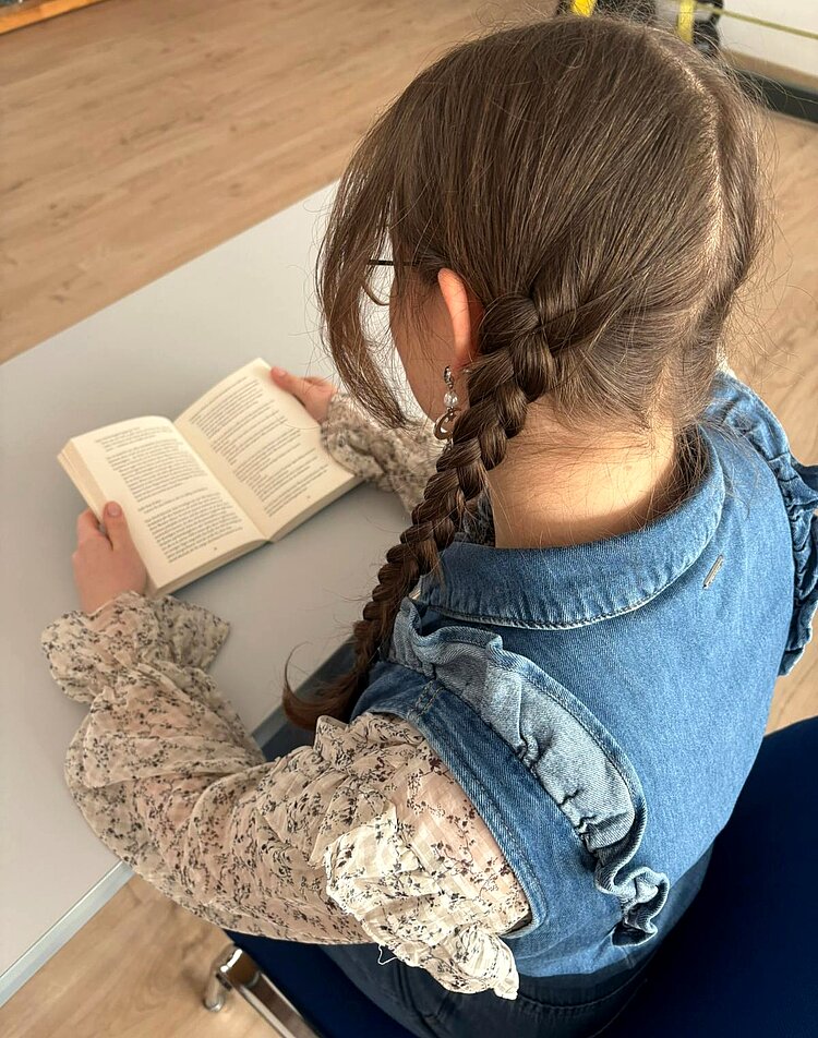 Von hinten aufgenommenes Mädchen mit geflochtenen Zöpfen und Brille, die ein aufgeschlagenes Buch hält.