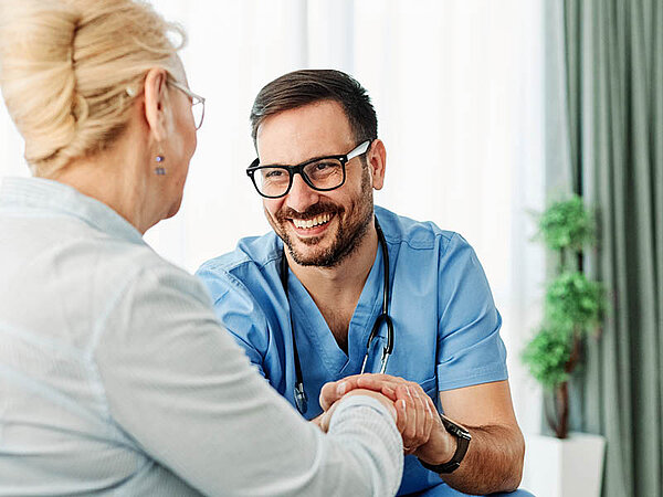 Ein Gesundheitsfachmann, erkennbar an den blauen OP-Kleidung und dem Stethoskop, hält die Hand einer anderen Person, deren Rücken zur Kamera zeigt. Die Szene spielt sich in einer Arztpraxis oder Klinik ab, wie durch Vorhänge und die saubere, professionelle Umgebung angedeutet. Der Fokus auf die Interaktion zwischen dem Gesundheitsfachmann und der Patientin weist auf einen Moment der Fürsorge oder Unterstützung hin, der in Diskussionen über Gesundheitsdienste, Patientenbetreuung oder medizinische Berufe relevant sein könnte.