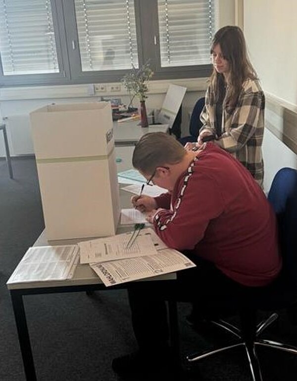 Testwahlen an der Euro Akademie Aschaffenburg am 19. Februar 2025 Ein Mann in einem roten Pullover sitzt an einem Tisch und füllt einen Stimmzettel aus, während eine junge Frau in kariertem Hemd hinter ihm steht. Eine Wahlkabine sorgt für Privatsphäre.