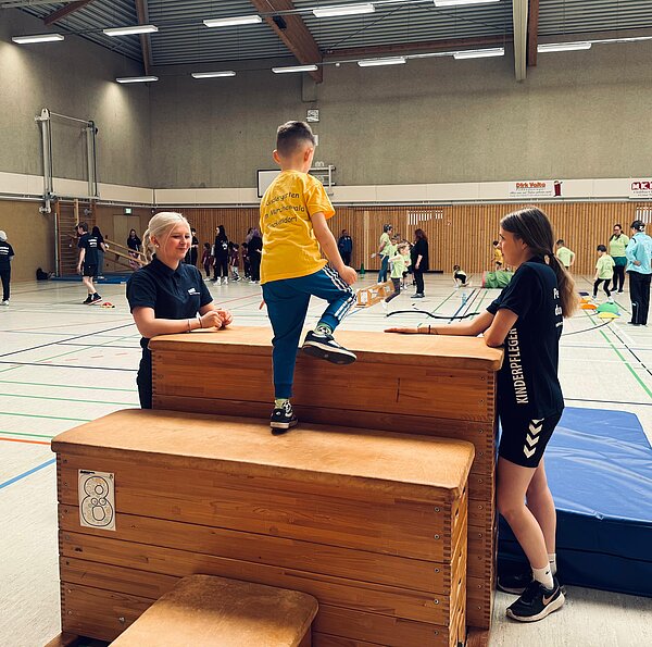 In einer Turnhalle klettert ein Junge in gelbem T-Shirt über zwei Holzböcke, assistiert von zwei Frauen in schwarzen T-Shirts; im Hintergrund sind weitere Kinder beim Sport zu sehen.