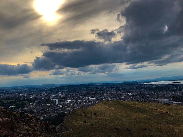 Aussicht von einem Berg auf eine Stadt, während am Himmel die Sonne durch Wolken diffus hindurchscheint und dunkle Wolken aufziehen. Im Vordergrund sind braune Felsen und weiter unten eine Grasfläche zu sehen.