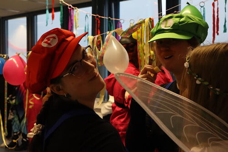 Fasching – Ein Tag voller Spaß und Gemeinschaft Zwei Schülerinnen, die als Mario und Luigi (von Super Mario Land) und demnach eine rote und eine grüne Mütze mit ihren Anfangsbuchstaben tragen, schauen für einen Schnappschuss in die Kamera. Im Hintergrund sieht man Luftballons und Luftschlange, vorne ins Bild drängt sich offenbar ein Engel, von dem man einen Flügel erkennen kann.