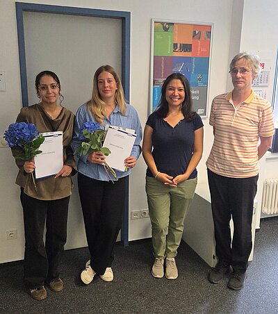 Vier Personen stehen nebeneinander in einem Büro: Zwei Frauen in der Mitte halten jeweils blaue Blumen und Urkunden, die anderen beiden stehen daneben und lächeln in die Kamera.