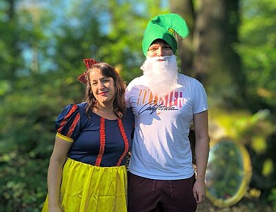 Eine Frau in einem Schneewittchen-Kostüm und ein Mann, der als ein Zwerg verkleidet ist mit Zipfelmütze und weißem Bart stehen einander umarmend in einer Waldumgebung.