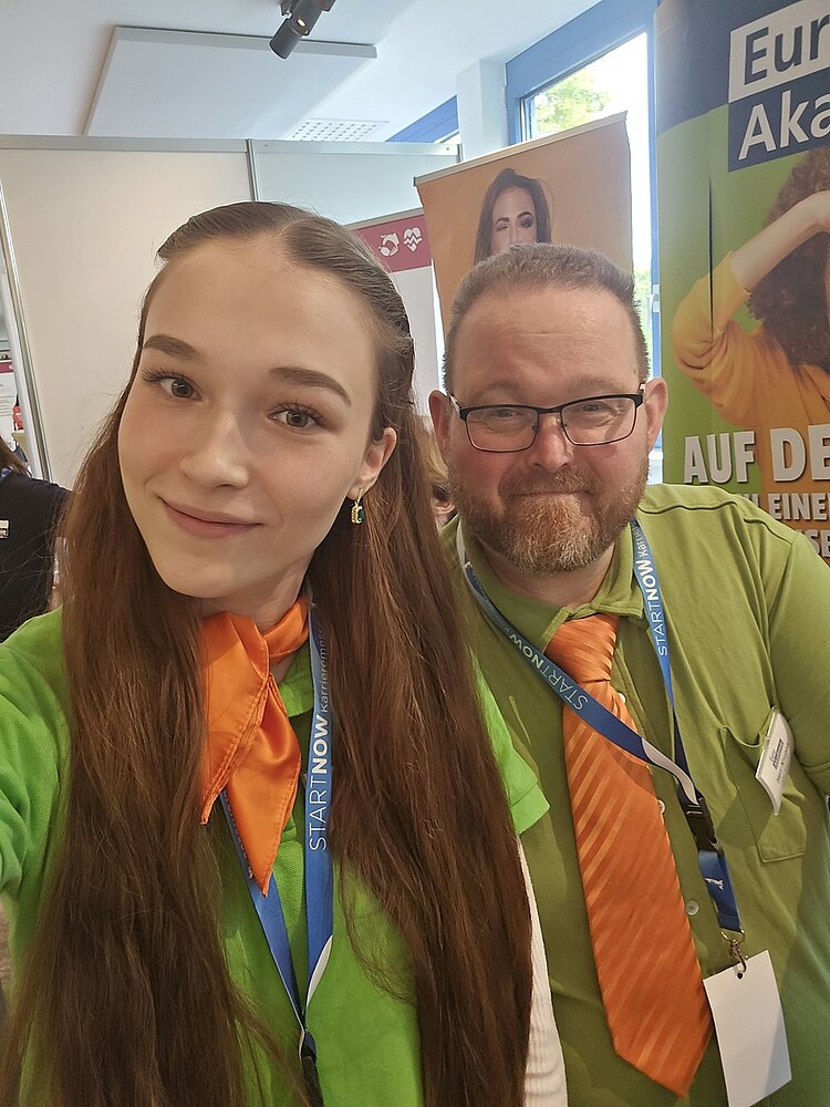 Die frisch gebackene Kauffrau für Büromanagement steht neben einem Kollegen der Euro Akademie Halle, beide blicken lächelnd in die Kamera. Beide tragen grüne Oberteile, orange Krawatten und blaue Namensbänder um den Hals. Im Hintergrund sind Roll-ups mit Werbemotiven und Texten zu sehen. Die Szene spielt in einem hellen Raum mit großen Fenstern.