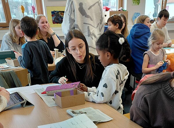 Eine junge Frau mit Brille sitzt an einem Tisch und erklärt einem Mädchen mit gepunktetem Hoodie ein lilafarbenes Kartonhaus; rundherum sitzen weitere Kinder und Jugendliche, die konzentriert an ihren Bastelprojekten arbeiten.
