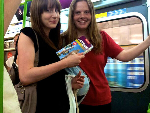 Zwei lächelnde junge Frauen in einer U-Bahn. Eine der Frauen hält einen Reiseführer von London hoch und trägt eine Handtasche.