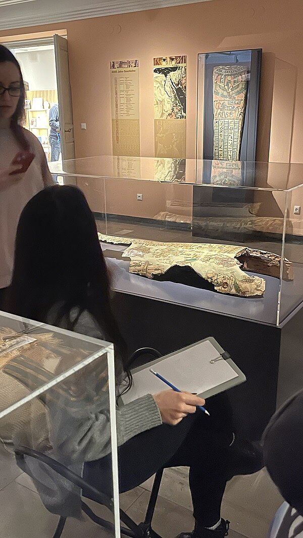 Zwei weibliche Auszubildende im Ausstellungsraum, eine sitzt und hält ihren Skizzenblock auf dem Schoß, die andere steht neben ihr.