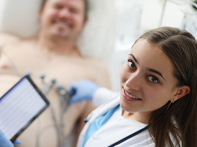 Auf dem Bild ist eine Ärztin mit einem Tablet zu sehen, während im Hintergrund ein Patient liegt, der am Herz untersucht wird.