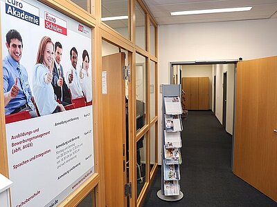 Blick in den langen Flur der Euro Akademie Dresden, links hängen zwei Poster der Euro Akademie und der Euro-Schulen Dresden, ein schmales Regal steht seitlich, in ihm stehen Informationsbroschüren und Flyer.