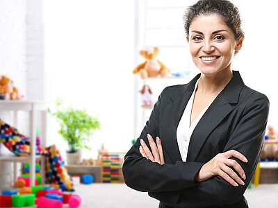 Porträt einer lächelnden Frau in einem schwarzen Blazer mit verschränkten Armen. Sie steht in einem hellen Raum, der wie eine Kindertagesstätte oder ein Spielzimmer eingerichtet ist – im Hintergrund sind bunte Spielzeuge, Plüschtiere und Regale zu sehen.