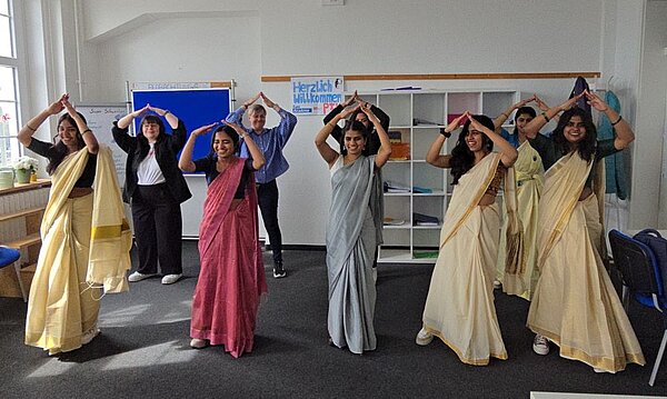 Eine gemischte Gruppe aus indischen Auszubildenden in traditioneller Kleidung und zwei deutschen Lehrkräften führt gemeinsam eine Tanzbewegung mit erhobenen Armen aus.