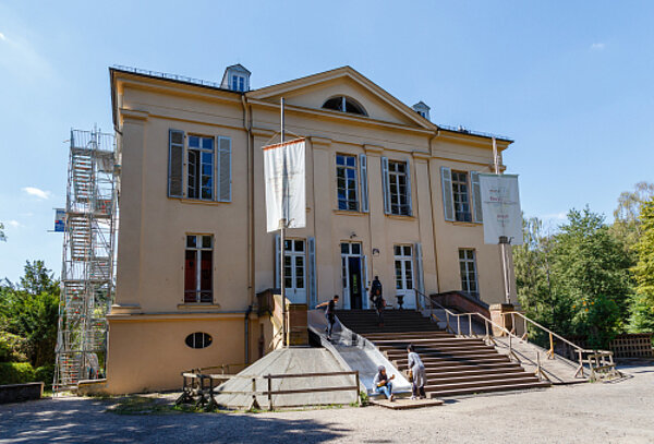Völlig neue Sinneswelten Ein in hellem gelb gestrichenes Museumsgebäude mit großen, geöffneten, hellblauen Fensterläden. Eine breite Treppe führt zum Eingang, links und rechts hängen Fahnen an einem Mast. Auf der linken Seite der Treppe führt eine Rutsche nach unten, auf der sich gerade eine stehende Person befindet.