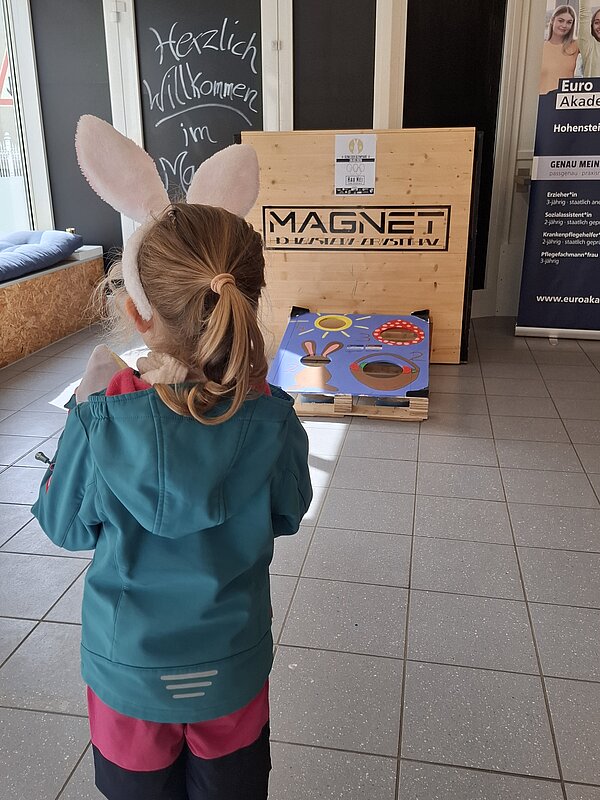 Ein Kind mit Hasenohren steht vor einem österlich bemalten Wurfspiel im Foyer der Euro Akademie.