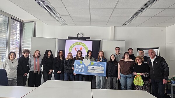 Die jungen Auszubildenden der Euro Akademie Erfurt stehen nebeneinander in einem Klassenraum. Im Hintergrund ist eine große Präsentationsfläche mit einem farbigen Logo und einem Schriftzug zu sehen. Zwei Azubis halten gemeinsam einen großen Check mit einem Herzsymbol und deutlich lesbarer Schrift. Die Fenster auf der linken Seite lassen Tageslicht herein. Die Kleidung der Personen ist unterschiedlich, von Freizeitkleidung bis zu Jacken.