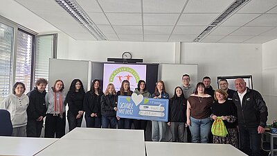 Die jungen Auszubildenden der Euro Akademie Erfurt stehen nebeneinander in einem Klassenraum. Im Hintergrund ist eine große Präsentationsfläche mit einem farbigen Logo und einem Schriftzug zu sehen. Zwei Azubis halten gemeinsam einen großen Check mit einem Herzsymbol und deutlich lesbarer Schrift. Die Fenster auf der linken Seite lassen Tageslicht herein. Die Kleidung der Personen ist unterschiedlich, von Freizeitkleidung bis zu Jacken.