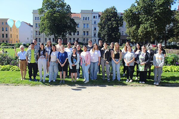Herzlich Willkommen zum ersten Schuljahr an der Euro Akademie Görlitz! Auf einer sonnigen Wiese inmitten der Stadt haben sich die neuen Auszubildenden für ein Gruppenfoto versammelt. Zwei hellblau Luftballons haben es auch auf das Bild geschafft.