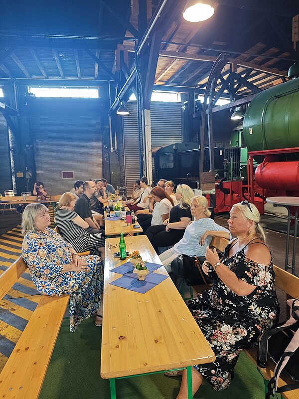 Eine Gruppe von Menschen sitzt an langen Holzbänken in einer großen, industriell gestalteten Halle. Die Halle ist mit Metallträgern und Oberlichtern ausgestattet. Im Hintergrund sind historische Züge und technische Geräte zu erkennen. Die Atmosphäre wirkt gesellig, und einige Personen unterhalten sich oder essen.
