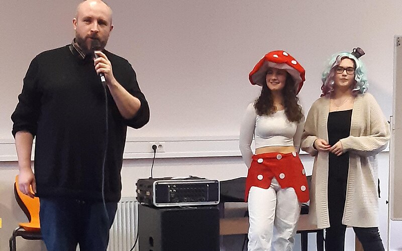 Drei Personen befinden sich in einem Raum und scheinen sich einem Publikum zuzuwenden. Ein Mann spricht in ein Mikrofon, während die anderen beiden Frauen in Kostümen gekleidet sind, eine als Pilz und die andere mit buntem Haar.