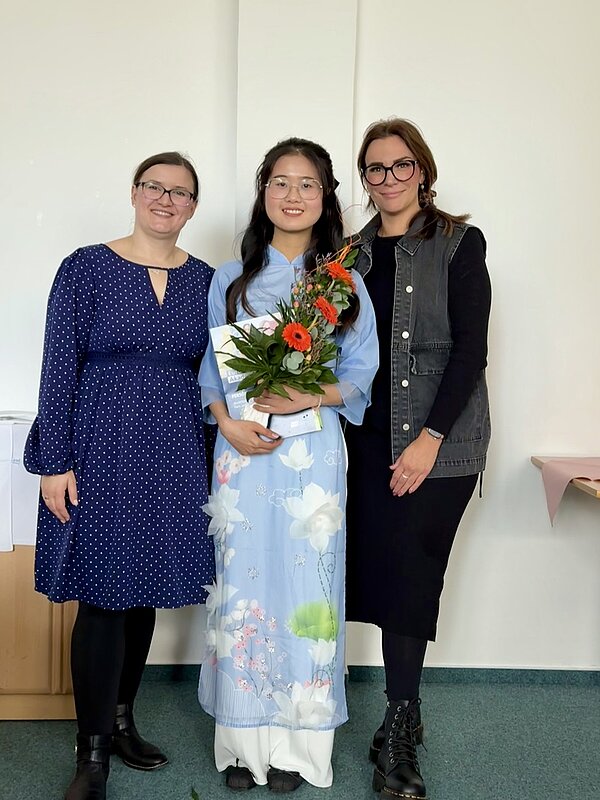 Eine Absolventin steht zwischen zwei ihrer Ausbilderinnen und posiert gemeinsam mit ihnen, anlässlich der Zeugnisübergabe an der Euro Akademie Bitterfeld-Wolfen, für ein Bild. Die Absolventin in der Mitte trägt ein hellblaues Kleid mit Blumenmuster und hält einen farbigen Blumenstrauß sowie ihr Zeugnis in den Händen.
