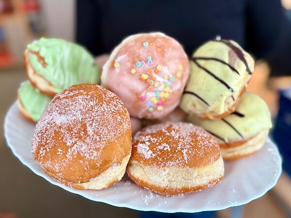 Teller mit verschiedenen Berlinern in bunten Glasuren und Zuckerguss, serviert zum Fasching am 11.11.2025 um 11:11 Uhr während einer Pause an den Euro-Schulen und den Euro Akademien Dresden, Meißen und Riesa. Die Süßspeisen schaffen, zusätzlich zur närrischen Jahreszeit, eine besondere Wohlfühlatmosphäre im Schulalltag.