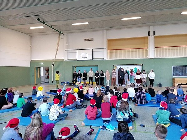 Engagiert und kreativ Weihnachtsaufführung auf der Bühne, alle Kinder sitzen auf Matten davor und verfolgen die Aufführung.