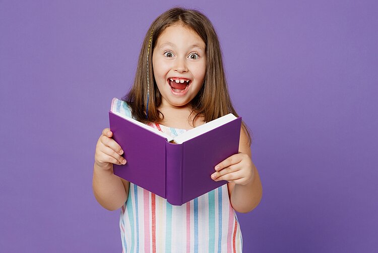 Kinderbuchklassiker im Unterricht kritisch beleuchtet · Teil 3 Ein kleines Mädchen in einem bunt gestreiften Kleid hält ein lila Buch in der Hand, das farblich mit dem Hintergrund harmoniert. Sie hat Augen und Mund weit aufgerissen, sodass man ihre Zahnlücken gut erkennt. In ihre glatten, braunen Haare ist eine bunte Strähne eingeflochten.