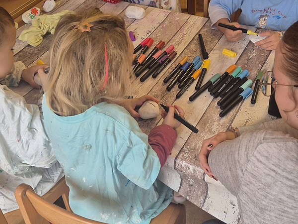 Kinder bemalen helle Steine mit Filzstiften an einem Basteltisch; eine erwachsene Person unterstützt.