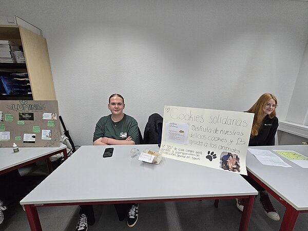 Drei Auszubildende sitzen an nebeneinanderstehenden Tischen in einem Raum mit weißen Wänden. Auf dem mittleren Tisch steht ein großes, handbeschriftetes Plakat mit dem Titel 'Cookies solidarias', darunter weitere spanische Schrift und eine gezeichnete Pfote. Auf dem Tisch liegen eine durchsichtige Plastikbox mit Keksen und einige Zettel. Links und rechts sind weitere Tische mit Plakaten und Papieren zu sehen. Die Personen blicken nach vorne oder zur Seite, tragen legere Kleidung und sitzen entspannt.