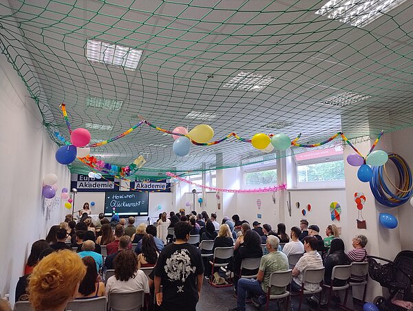 Woche der feierlichen Verabschiedungen Ein großer Raum mit einer Menschenmenge, die auf Stühlen sitzt und einer Veranstaltung zuhört. Im Vordergrund sind bunte Luftballons und Girlanden zu sehen, die an der Decke hängen. Im Hintergrund steht eine Bühne mit einer Rednerin vor einem Banner mit der Aufschrift “Euro Akademie”. Das Bild vermittelt eine festliche Atmosphäre bei einer offiziellen Veranstaltung oder Feier.
