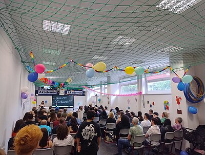  Ein großer Raum mit einer Menschenmenge, die auf Stühlen sitzt und einer Veranstaltung zuhört. Im Vordergrund sind bunte Luftballons und Girlanden zu sehen, die an der Decke hängen. Im Hintergrund steht eine Bühne mit einer Rednerin vor einem Banner mit der Aufschrift “Euro Akademie”. Das Bild vermittelt eine festliche Atmosphäre bei einer offiziellen Veranstaltung oder Feier.