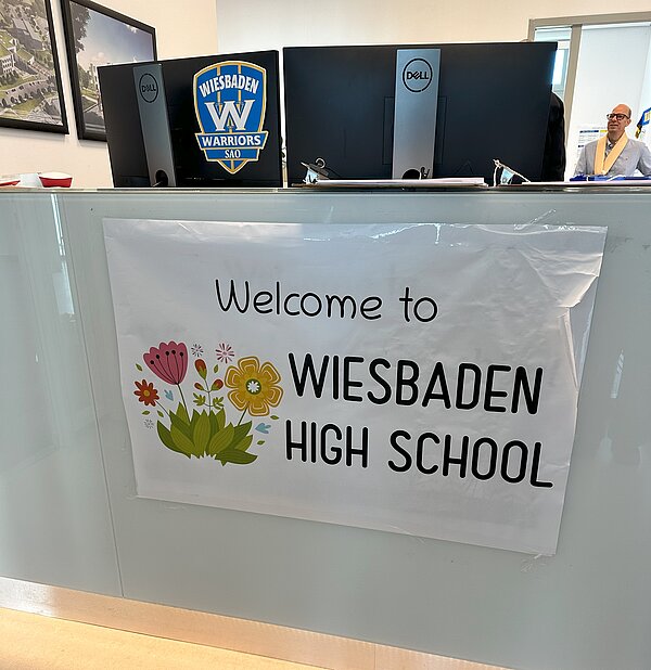 Empfangsbereich der Wiesbadener High School mit einem Willkommensschild, das 'Welcome to Wiesbadener High School' zeigt. Im Hintergrund sind ein Computerbildschirm mit dem Logo der 'Wiesbaden Warriors SAO' und ein Mann am Schalter zu sehen.