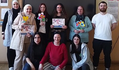 Eine Gruppe von Menschen, darunter Frauen mit Kopftüchern, posiert für ein Foto vor einer Tafel. Einige halten bunte Papierblumen und Flyer in den Händen. Die Gruppe lächelt und zeigt stolz ihre kreativen Werke.