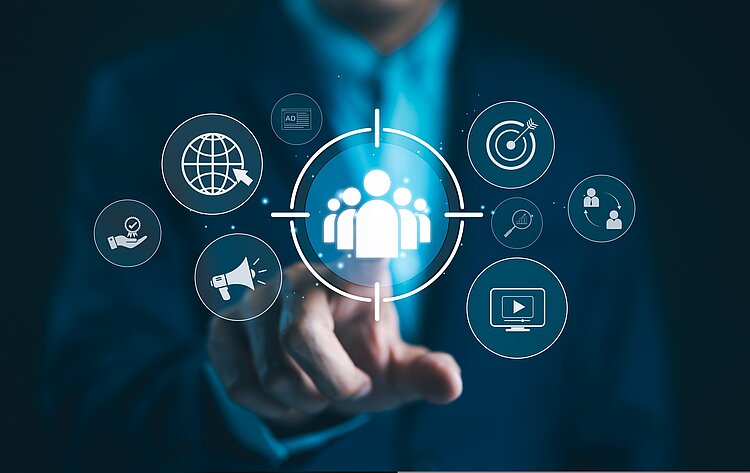 Eine Person in einem dunklen Anzug zeigt mit dem Finger auf ein holografisches Symbol in der Mitte des Bildes, das eine Gruppe von Menschen innerhalb eines Fadenkreuzes darstellt. Rundherum schweben verschiedene digitale Icons, darunter ein Globus, eine Zielscheibe mit einem Pfeil, eine Lupe mit Diagramm, ein Megafon, ein Video-Symbol und eine Hand mit einem Qualitätssiegel. Das Bild symbolisiert digitales Marketing, Zielgruppenanalyse und strategische Kundenansprache.