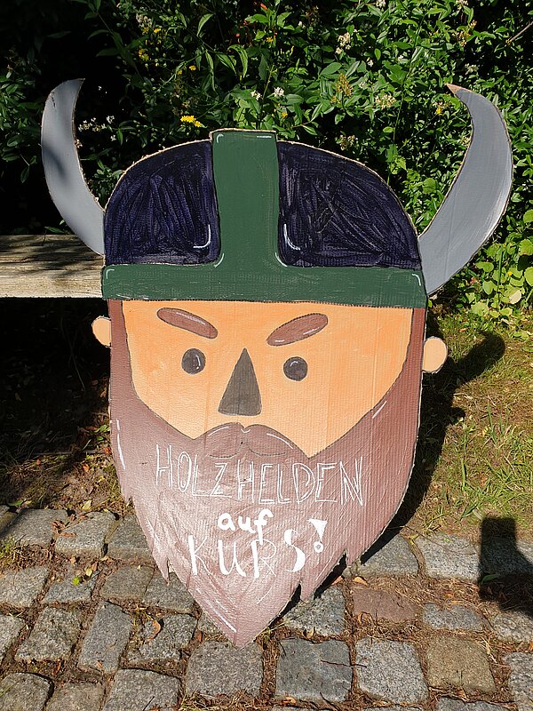 Ein erlebnisreicher Vormittag voller Spiel, Kreativität und Gemeinschaft Ein bemaltes Schild mit einem Wikingerkopf und der Aufschrift "Holzhelden auf Kurs" auf einem gepflasterten Weg vor grünen Büschen.