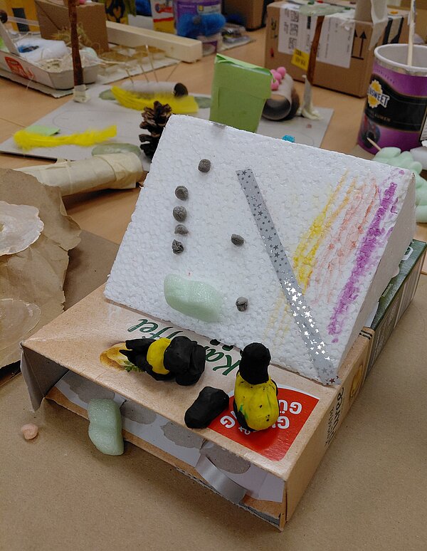 Aus einer flachen Schachtel und einer schräg aufgeklebten Styroporplatte ist ein Modell gebaut, das mit kleinen Steinen, Glitzerstreifen, Farbstreifen und geformten Knetfiguren verziert ist; im Hintergrund sind weitere Bastelarbeiten zu sehen.