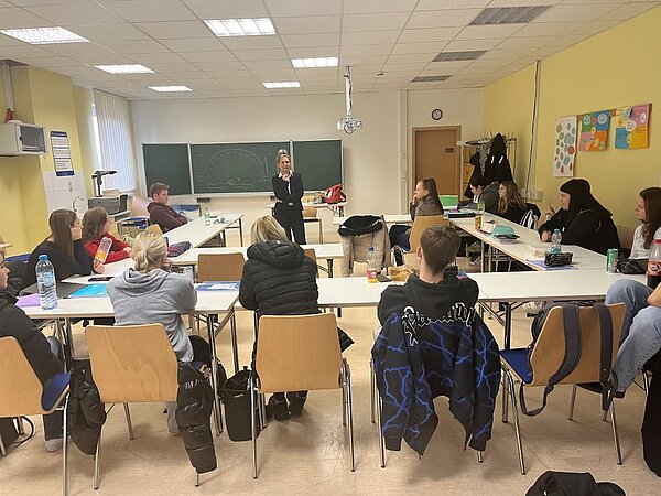 Eine Gruppe junger Erwachsener sitzt an langen Tischen in einem Klassenzimmer und hört einer stehenden Person zu, die vorne im Raum spricht. Die sprechende Person steht nahe der Tafel und scheint zu erklären oder zu unterrichten. Die Zuhörenden sind aufmerksam und blicken in ihre Richtung. Im Raum sind eine Tafel, ein Projektor und bunte Poster an der Wand zu sehen. Die Atmosphäre ist interaktiv und lernfördernd.